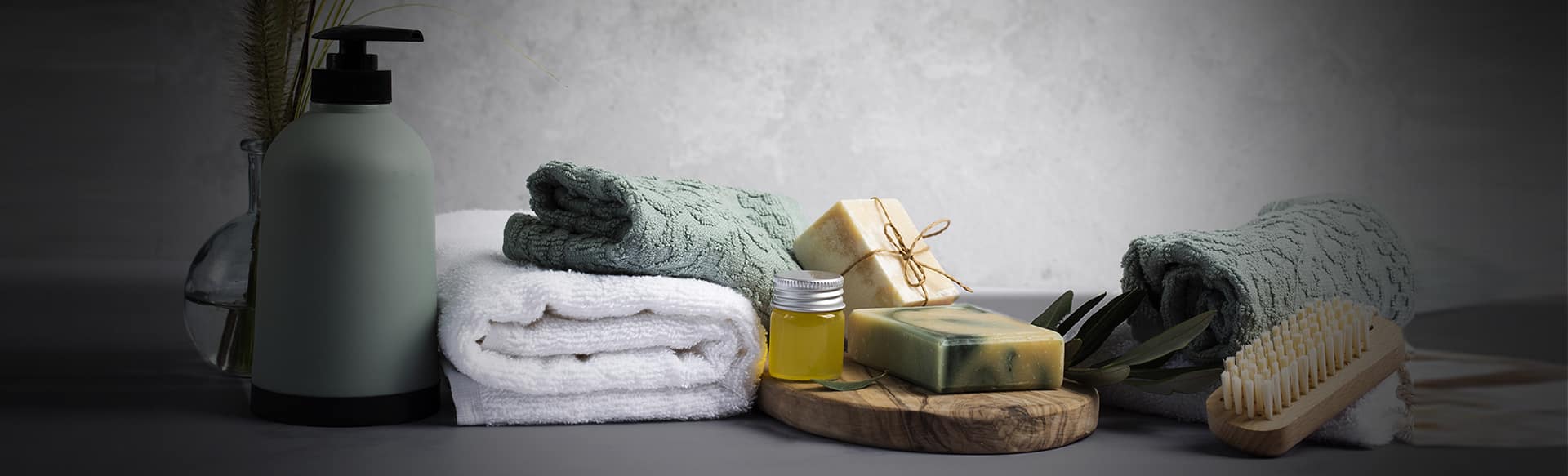 A spa-themed arrangement with folded green and white towels, a pump bottle, bars of soap, a small jar of yellow oil, a wooden brush, and a few green leaves, all set against a gray background. The setting exudes a calm and relaxing atmosphere.