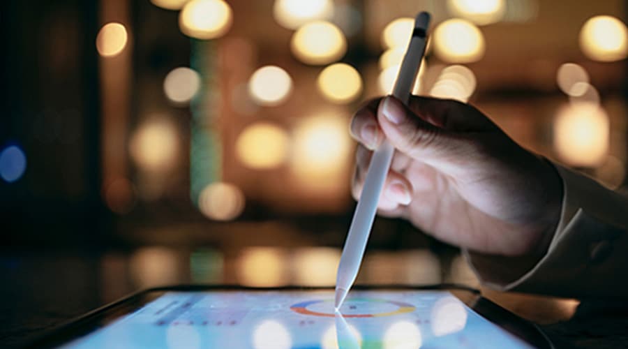 A person uses a stylus to interact with a tablet. The background is blurred, with warm, circular lights creating a soft bokeh effect, adding a cozy and focused atmosphere to the scene.