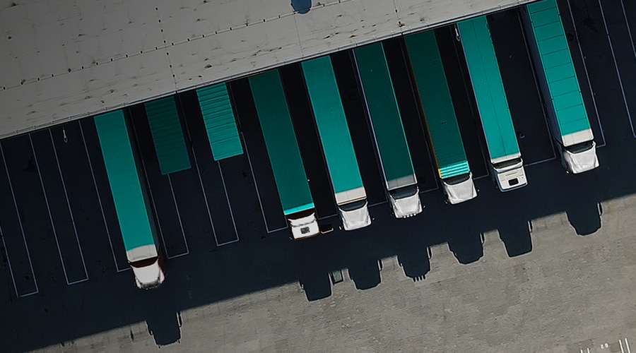 Aerial view of several semi-trucks parked in parallel at loading docks. The top of the image shows the roof of the warehouse with shadows cast on the pavement below. The trucks have green roofs and are lined up neatly in designated parking spaces.