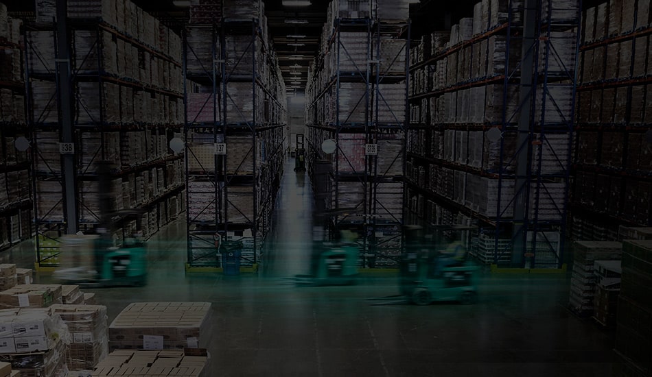 A large warehouse filled with tall shelving units stocked with various goods. Two forklifts, moving rapidly, appear as green blurs in the foreground. The central aisle extends into the distance, lined with well-organized pallets and boxes. The lighting is dim.