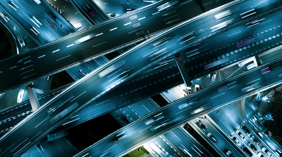 Aerial view of multiple illuminated highways and overpasses at night, with blurred headlights and taillights from moving vehicles creating light trails.