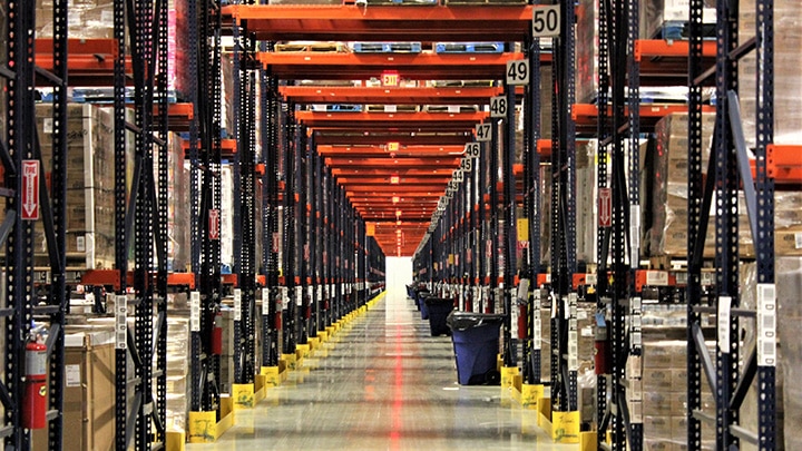 Central warehouse aisle with tall metal shelving, organized pallets, boxes, bright lighting, and a spotless shiny floor.