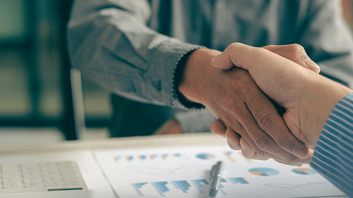 Two people shaking hands over a desk with financial charts and documents, indicating a successful business partnership or agreement.