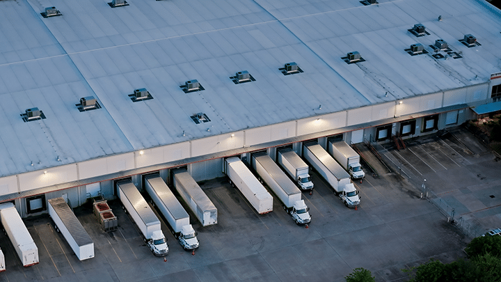 Aerial view of warehouse with blue-gray roof, white trucks at loading docks, and empty parking spaces nearby.
