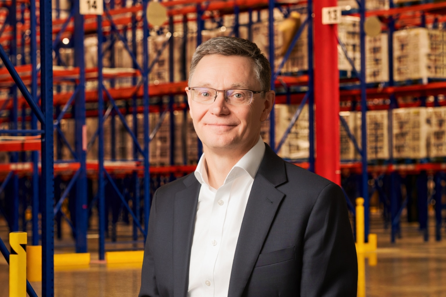 Boubacar Diallo in a dark suit and white shirt stands in a warehouse with tall shelves stocked with boxes in the background.