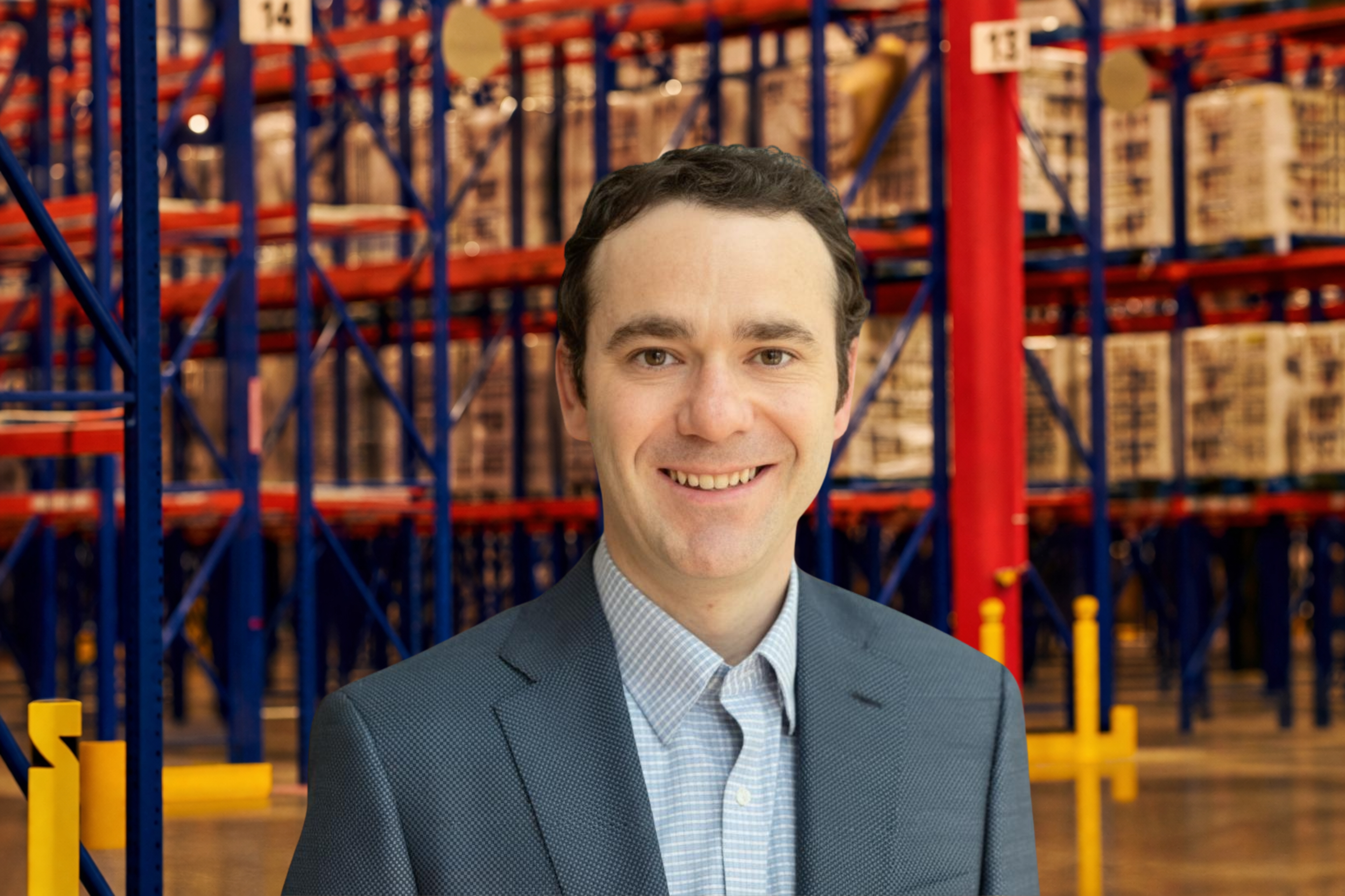 Spencer Smith in a blue suit and checked shirt smiles in a warehouse aisle with tall shelves filled with goods.