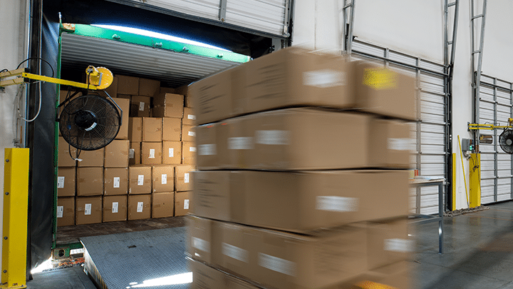 Cardboard boxes on a pallet are moved into a warehouse loading dock, with more boxes stacked inside a nearby truck trailer.