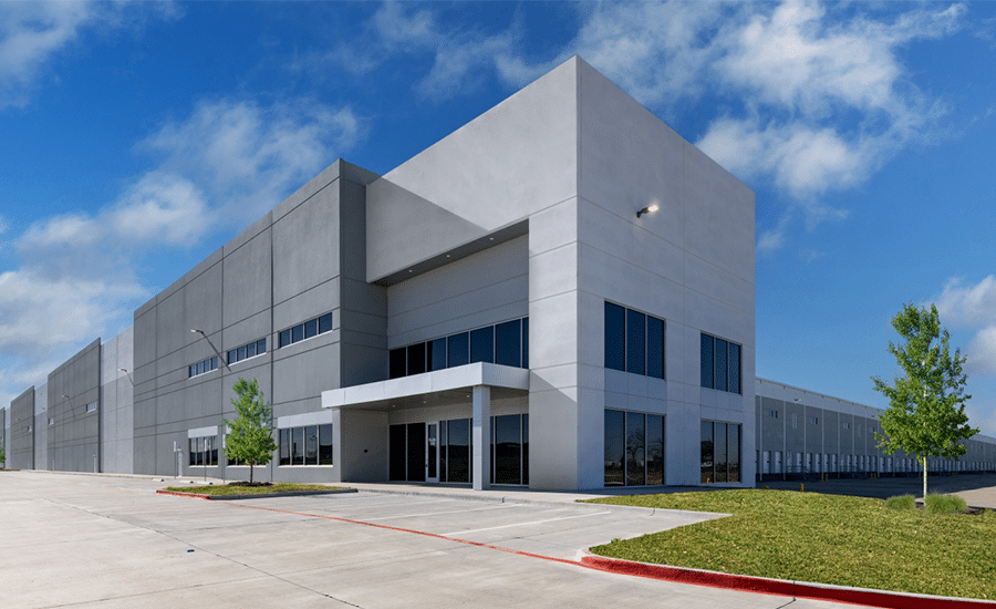 Modern gray industrial warehouse with tall windows, flat roof, empty concrete lot, small trees, and blue sky with clouds.